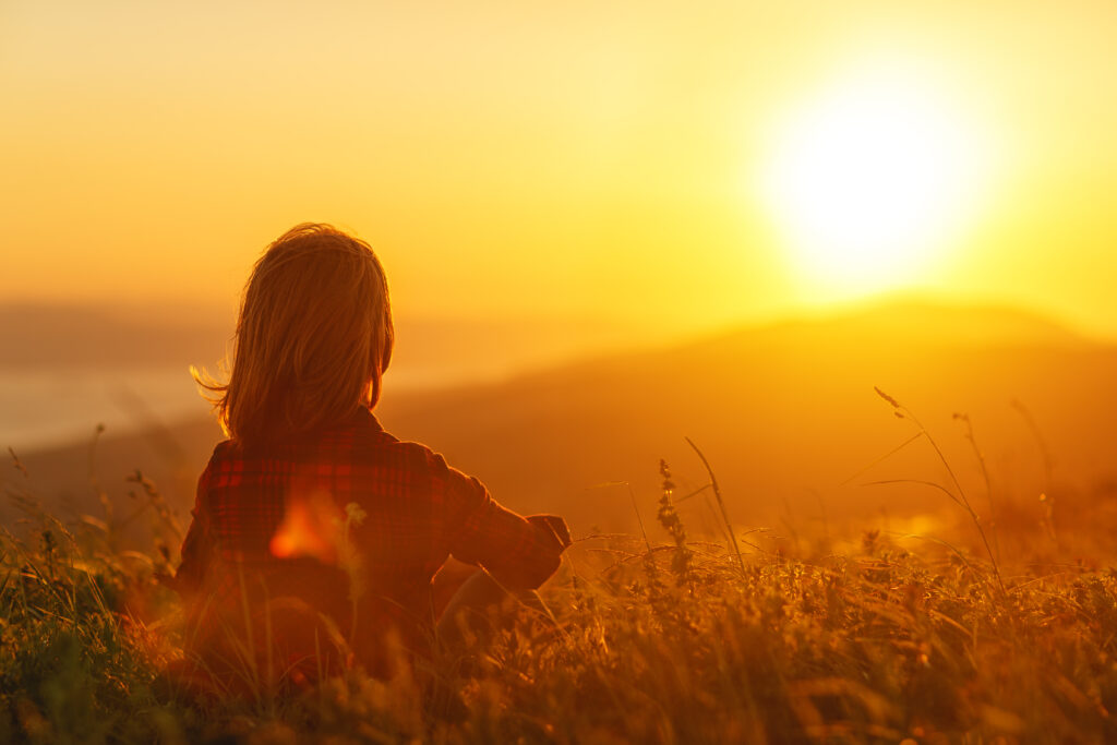 Woman sits with her back in the field and look sunset in the mountains
